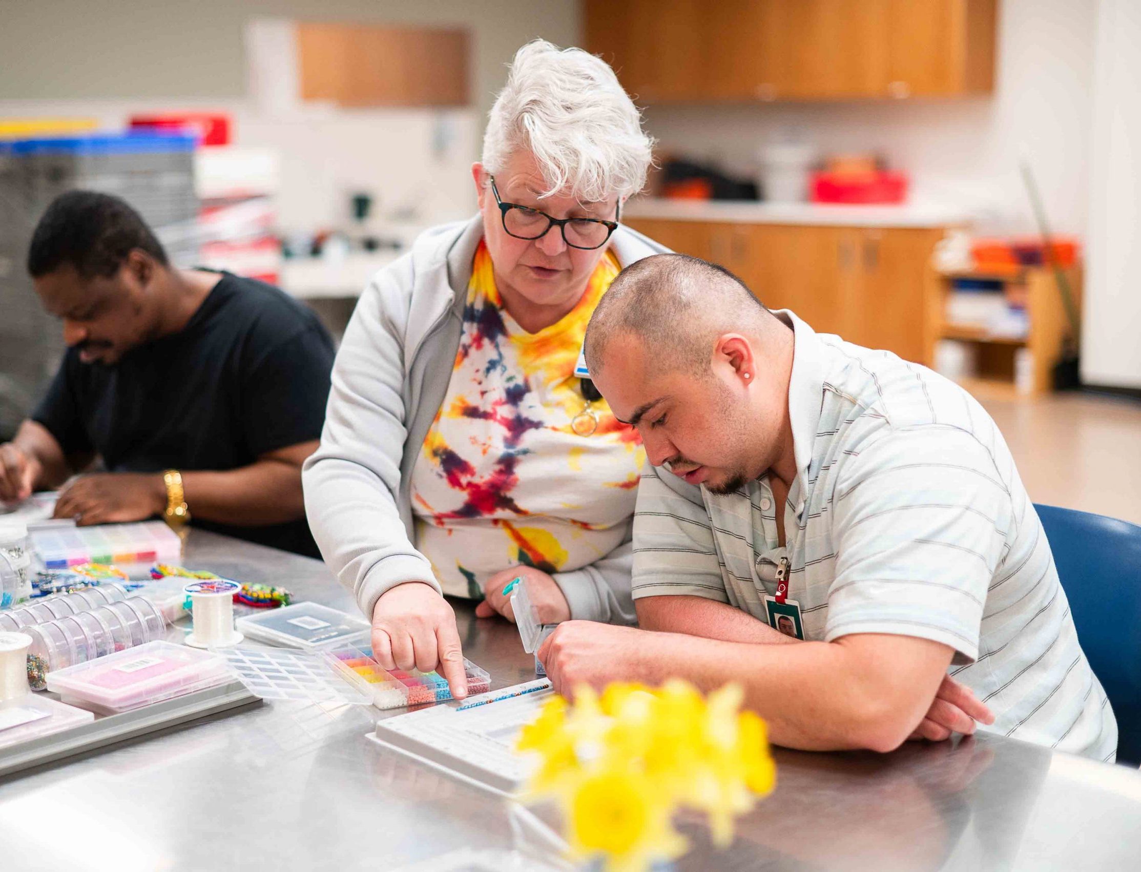 An instructor helps a seated man with an activity at a table.