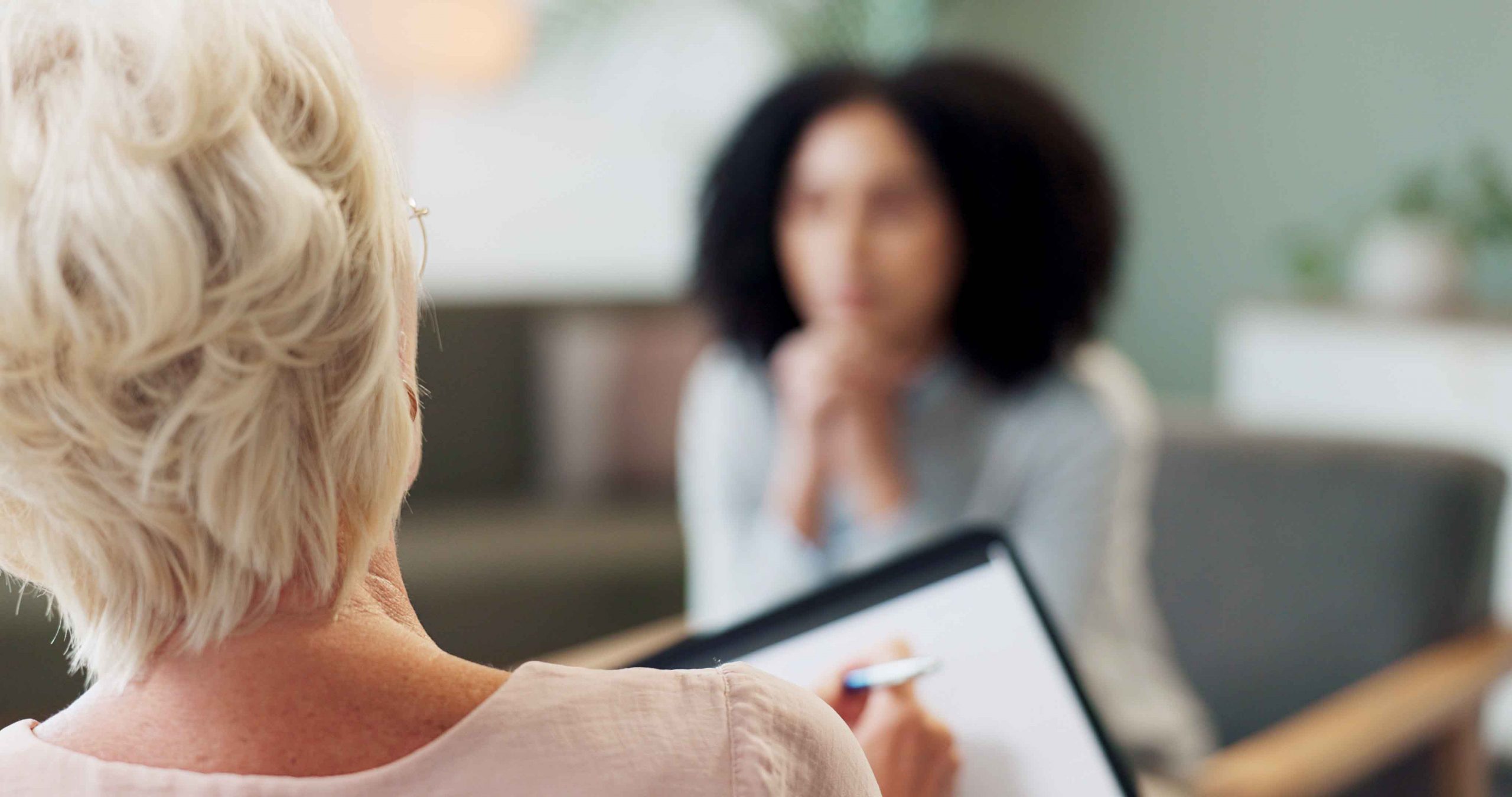 A therapist holds a clipboard and pen, facing a woman who appears to be speaking.