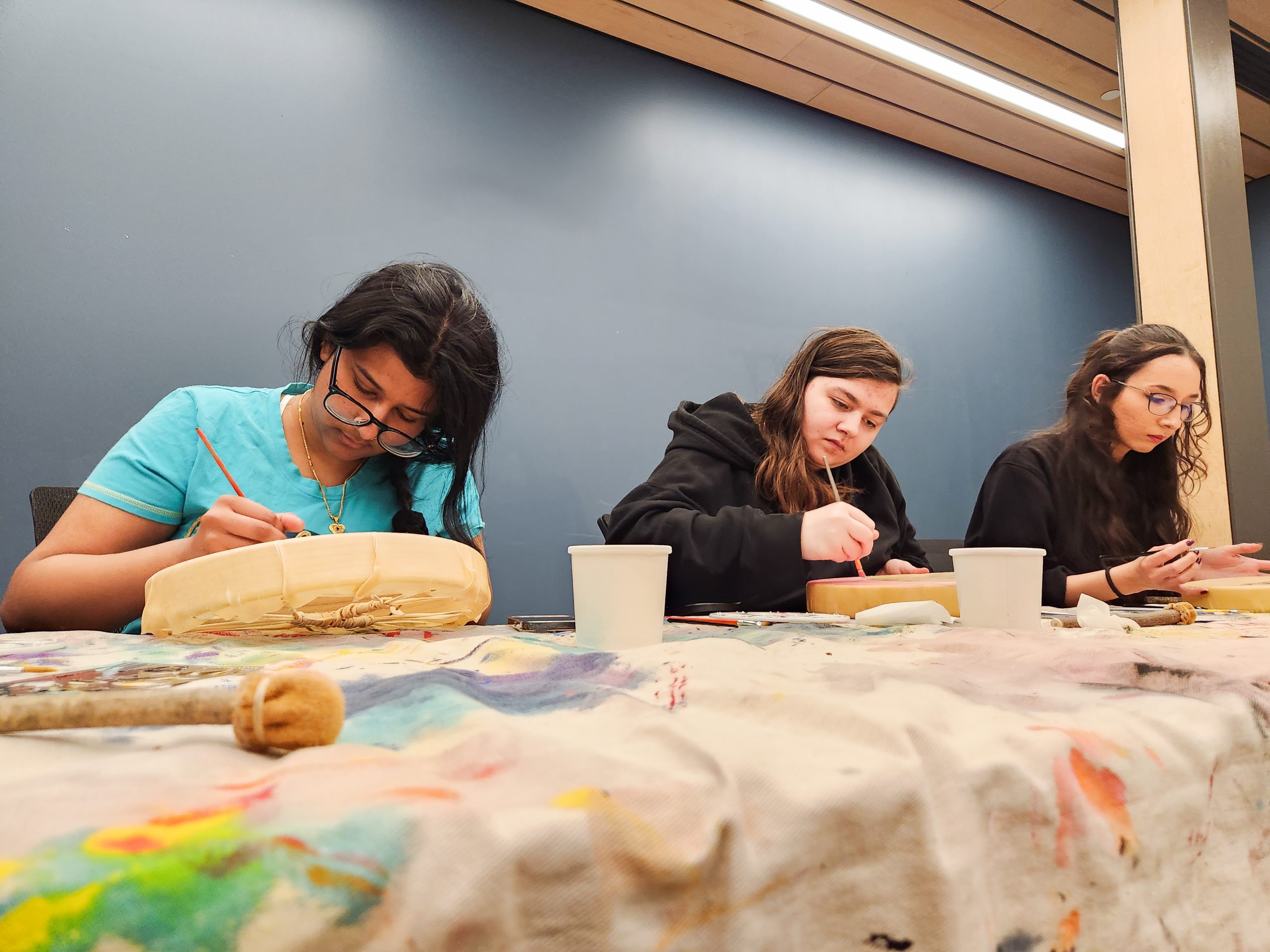 Three women sitting at a paint-splattered table, painting on round canvases.